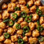 Close-up of a sizzling jalapeno chicken dish with golden-brown, bite-sized chicken pieces coated in a rich, glossy sauce, garnished with fresh jalapeño slices and sesame seeds, served in a skillet.