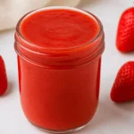 Homemade strawberry puree in a glass jar surrounded by fresh strawberries on a white surface.
