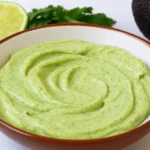 A bowl of creamy avocado crema with lime and cilantro in the background.