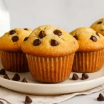 Freshly baked chocolate chip muffins on a plate with scattered chocolate chips and a bottle of milk in the background.