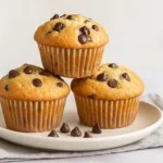 Freshly baked chocolate chip muffins stacked on a white plate, topped with melted chocolate chips.
