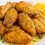 Close-up of crispy lemon pepper chicken wings on a white plate, garnished with chopped herbs and lemon slices.