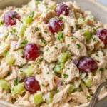 A close-up of creamy chicken salad with red grapes, celery, and herbs, served in a rustic ceramic bowl with a spoon.