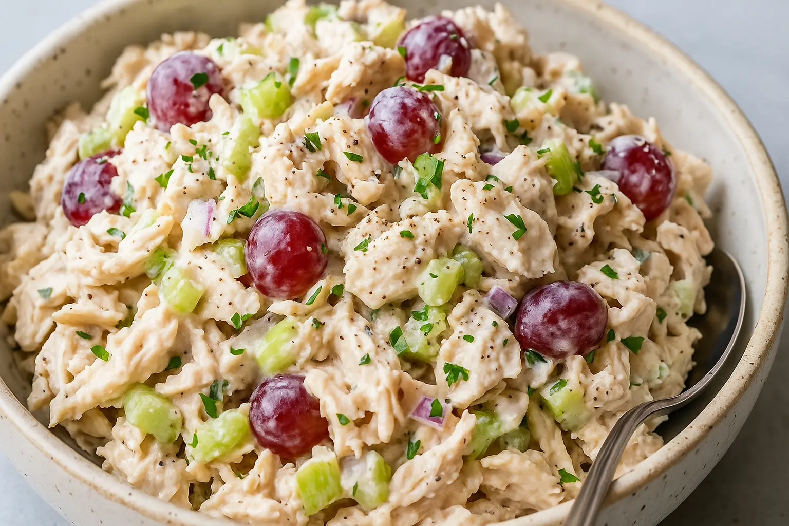 A close-up of creamy chicken salad with red grapes, celery, and herbs, served in a rustic ceramic bowl with a spoon.