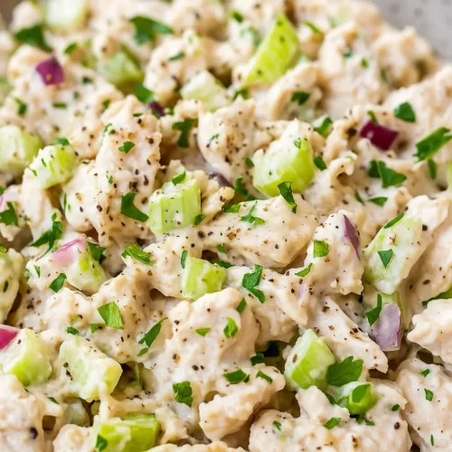 Close-up of creamy chicken salad with chopped celery, red onion, fresh parsley, and black pepper.