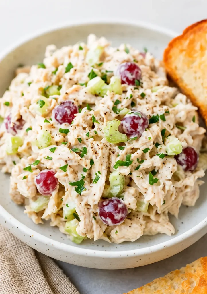 Plate of creamy chicken salad with red grapes, celery, and fresh herbs, served with toasted bread on the side.