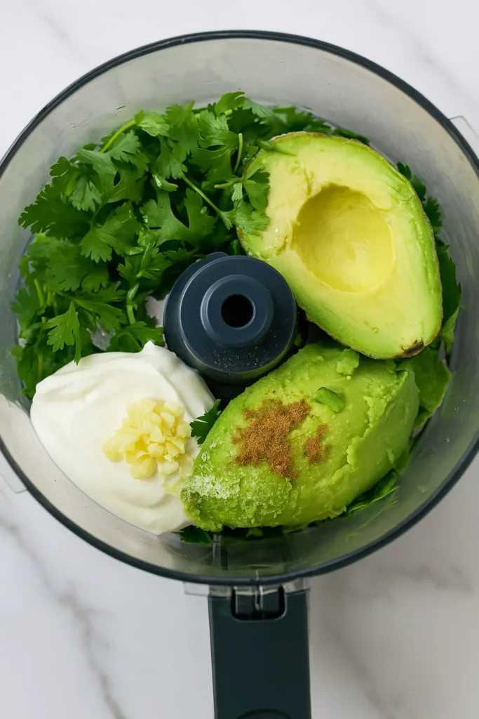 Fresh avocado, cilantro, yogurt, garlic, and cumin in a food processor bowl for avocado crema.