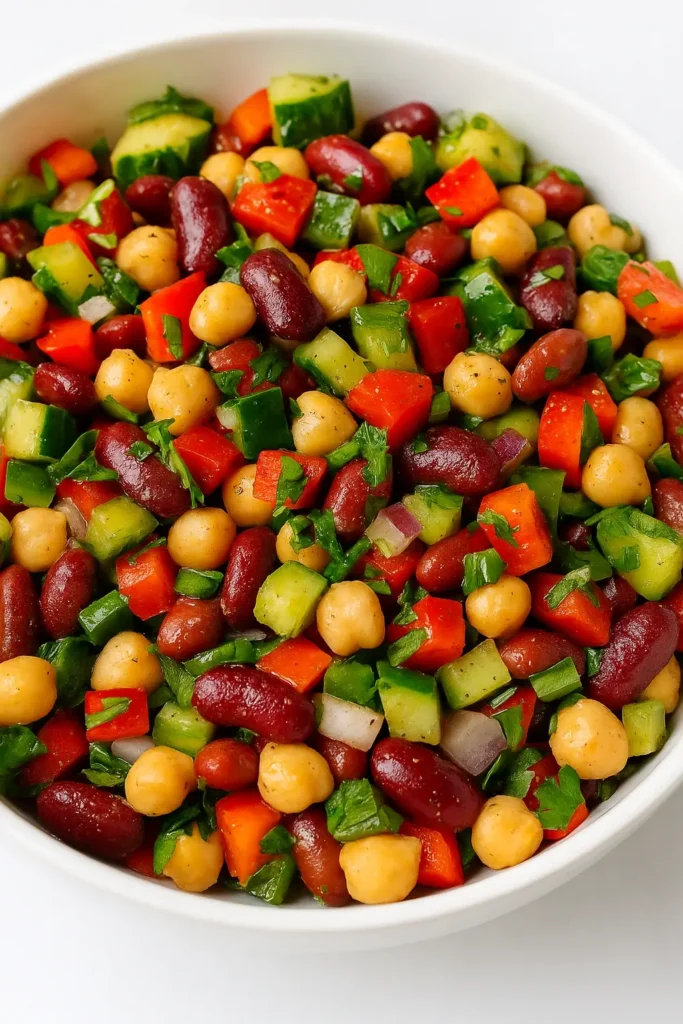 Overhead view of a vibrant dense bean salad with chickpeas, kidney beans, diced cucumbers, red bell peppers, and parsley in a white bowl.