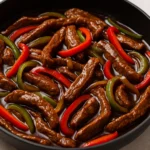 Sliced beef strips with red and green bell peppers in a glossy stir-fry sauce, served hot in a black skillet.